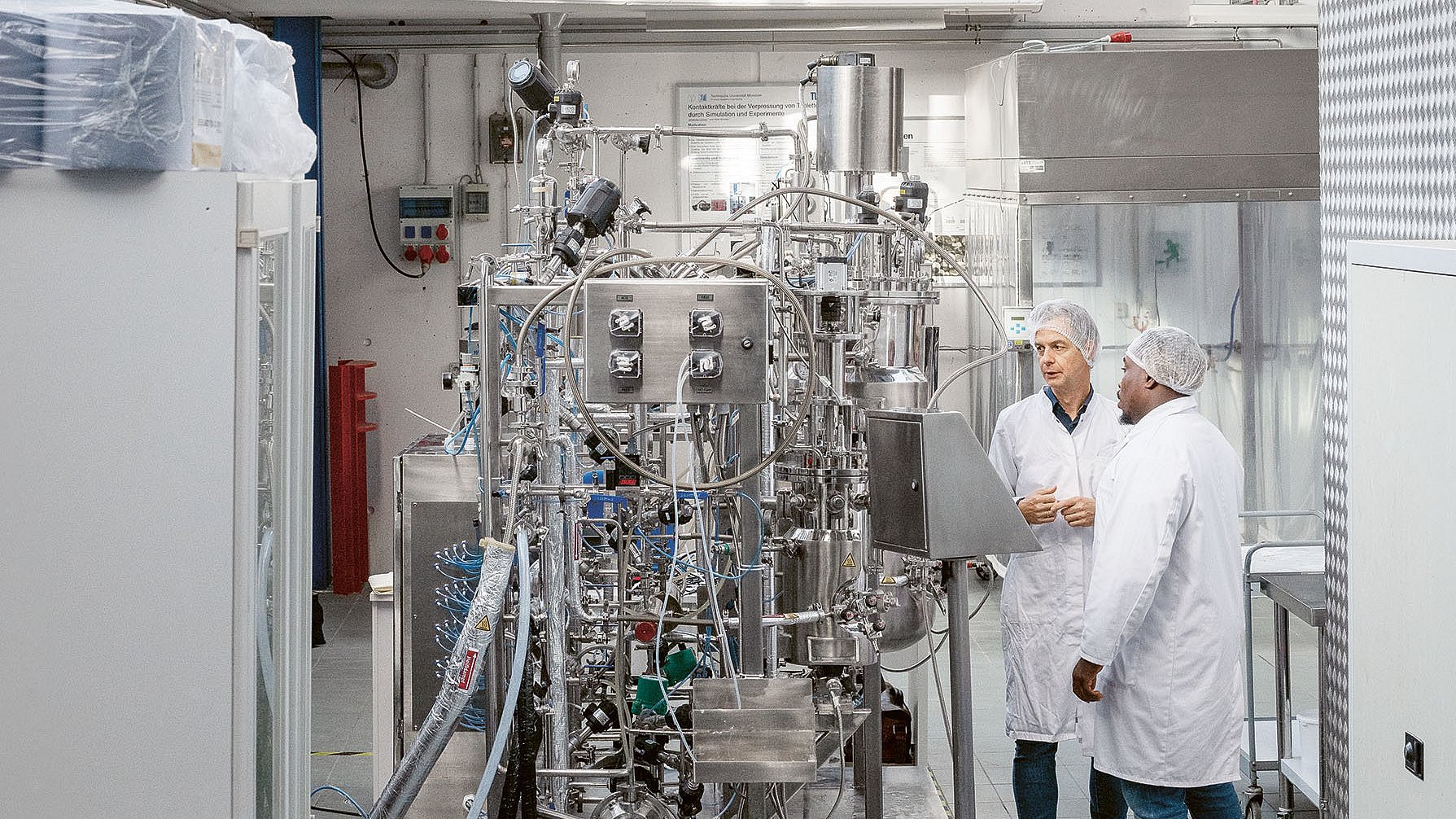  Das Bild zeigt zwei Männer neben einem Fermenter stehen. Sie tragen weiße Kittel und Haarnetze.  Sie stehen vor einer großen, komplizierten Maschine aus poliertem Edelstahl. Die Maschine ist mit vielen Rohren, Ventilen und Messgeräten ausgestattet und scheint ein komplexer chemischer oder biologischer Reaktor zu sein.  Links im Bild stehen mehrere weiße Schränke, die wahrscheinlich als Lager dienen.  Auf dem Boden sind Schläuche und Kabel zu sehen, die mit der Maschine verbunden sind. Die Atmosphäre ist sachlich, konzentriert und professionell. 