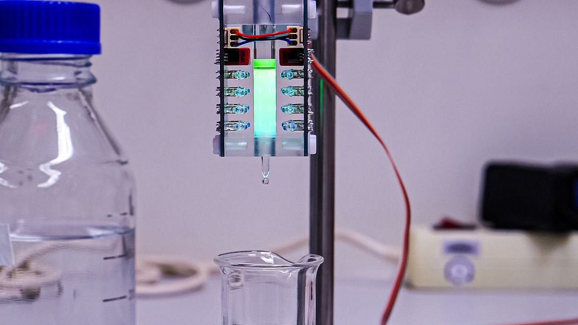 Das Bild zeigt die Apparatur, die das Team zur Arbeit im Labormaßstab entwickelt hat. Man sieht ein kleines Behältnis, das sich zwischen LED-Leuchten befindet. Die Flüssigkeit in dem Behältnis leuchtet leicht.