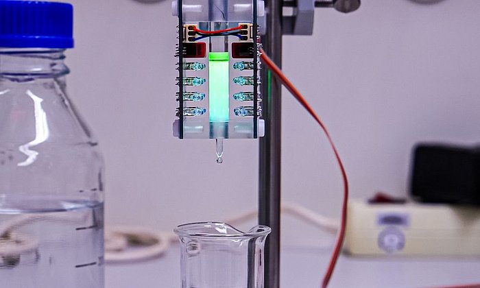 Das Bild zeigt die Apparatur, die das Team zur Arbeit im Labormaßstab entwickelt hat. Man sieht ein kleines Behältnis, das sich zwischen LED-Leuchten befindet. Die Flüssigkeit in dem Behältnis leuchtet leicht.