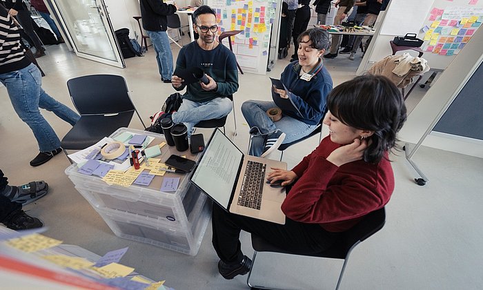 Three students at an improvised table made of two plastic containers, in the background partitions with a multitude of written-on colored sticky notes.