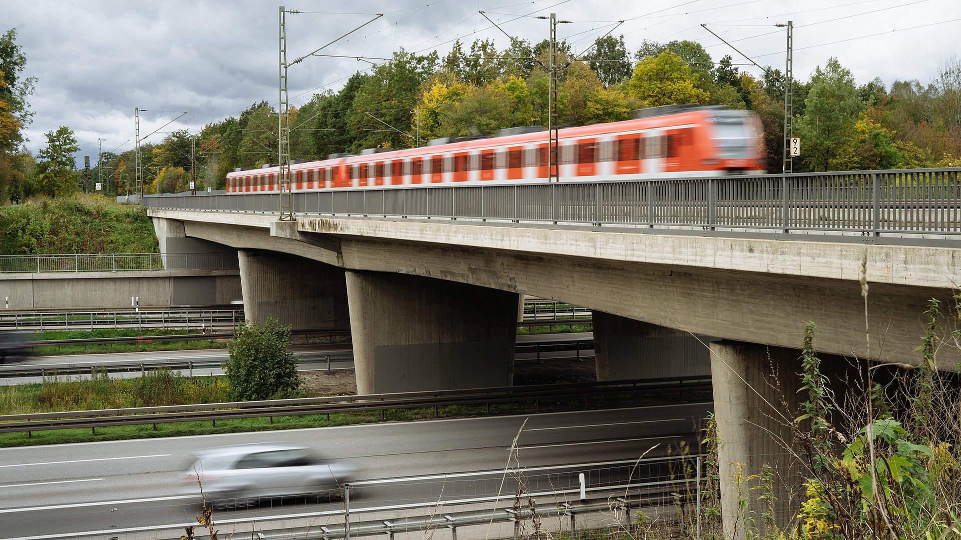 Photo shows a highway bridge on which a train is passing, a car is driving underneath