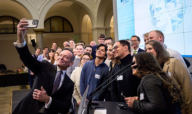 Bundesminister für Digitales und Verkehr Volker Wissing nimmt im Raum der Preisverleihung im Ministerium mit den Finalistinnen und Finalisten  der Digital Future Challenge 2025 ein Selfie auf; im Vordergrund ein Stehpult mit Mikrofonen, im Hintergrund rechts eine Leinwand mit projizierter Folie des Bundesministeriums für Digitales und Verkehr 