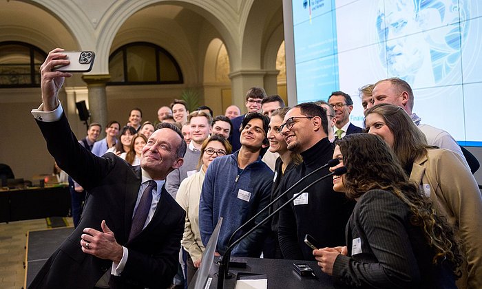 Bundesminister für Digitales und Verkehr Volker Wissing nimmt im Raum der Preisverleihung im Ministerium mit den Finalistinnen und Finalisten  der Digital Future Challenge 2025 ein Selfie auf; im Vordergrund ein Stehpult mit Mikrofonen, im Hintergrund rechts eine Leinwand mit projizierter Folie des Bundesministeriums für Digitales und Verkehr 