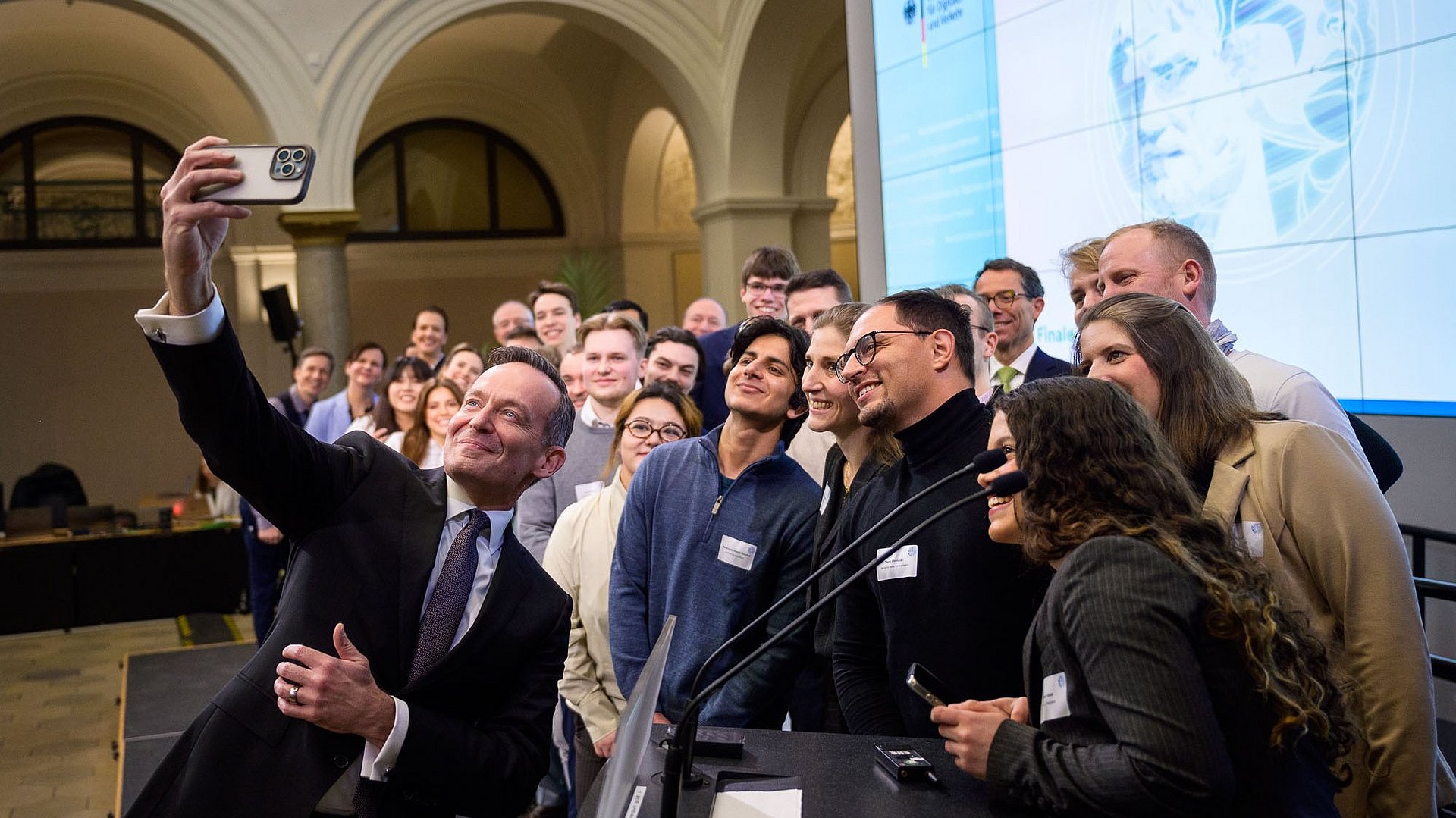 Bundesminister für Digitales und Verkehr Volker Wissing nimmt im Raum der Preisverleihung im Ministerium mit den Finalistinnen und Finalisten  der Digital Future Challenge 2025 ein Selfie auf; im Vordergrund ein Stehpult mit Mikrofonen, im Hintergrund rechts eine Leinwand mit projizierter Folie des Bundesministeriums für Digitales und Verkehr 