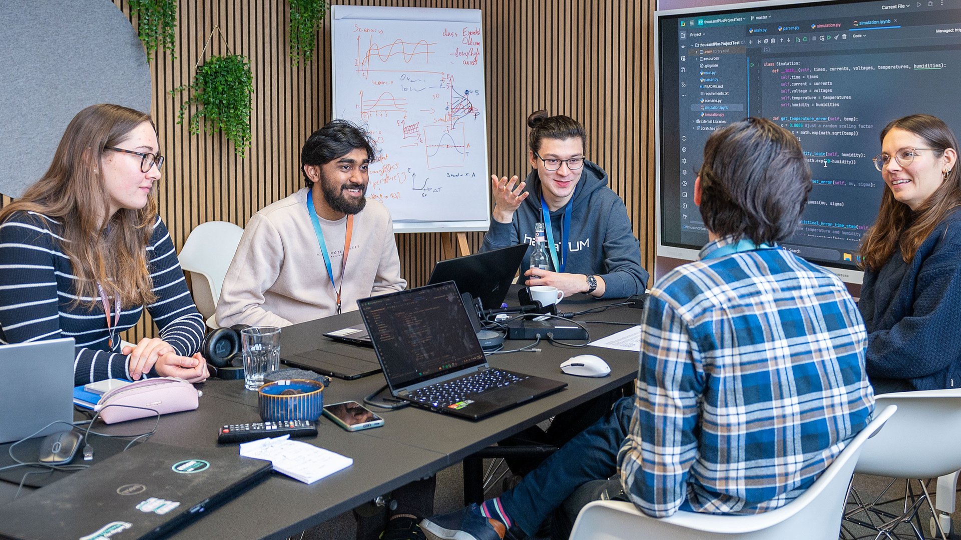 Fünf Studierende sitzen um einen Tisch und diskutieren. Vor zwei Studierenden steht ein Laptop. Sowohl auf den Laptops als auch auf dem Bildschirm im Hintergrund ist Quellcode zu sehen.