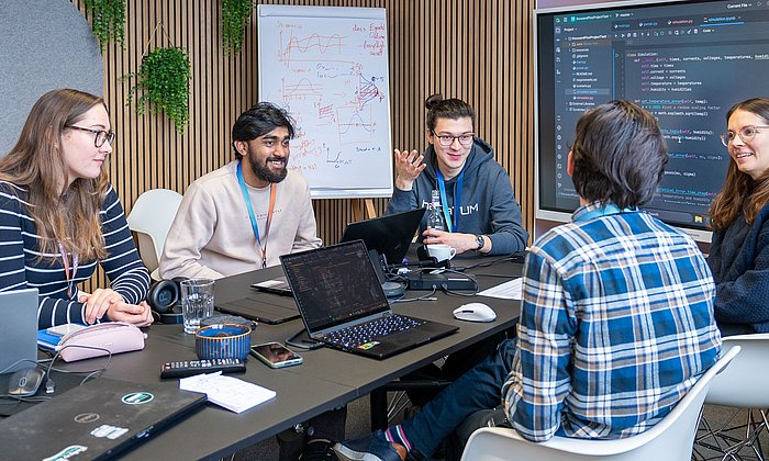 Five students sit around a table and discuss. There is a laptop in front of two students. Source code can be seen both on the laptops and on the screen in the background.