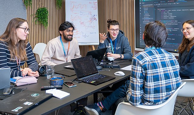 Fünf Studierende sitzen um einen Tisch und diskutieren. Vor zwei Studierenden steht ein Laptop. Sowohl auf den Laptops als auch auf dem Bildschirm im Hintergrund ist Quellcode zu sehen.