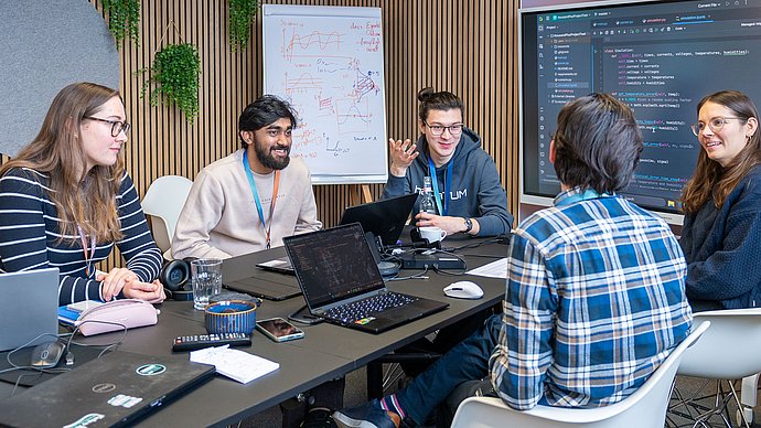 Five students sit around a table and discuss. There is a laptop in front of two students. Source code can be seen both on the laptops and on the screen in the background.