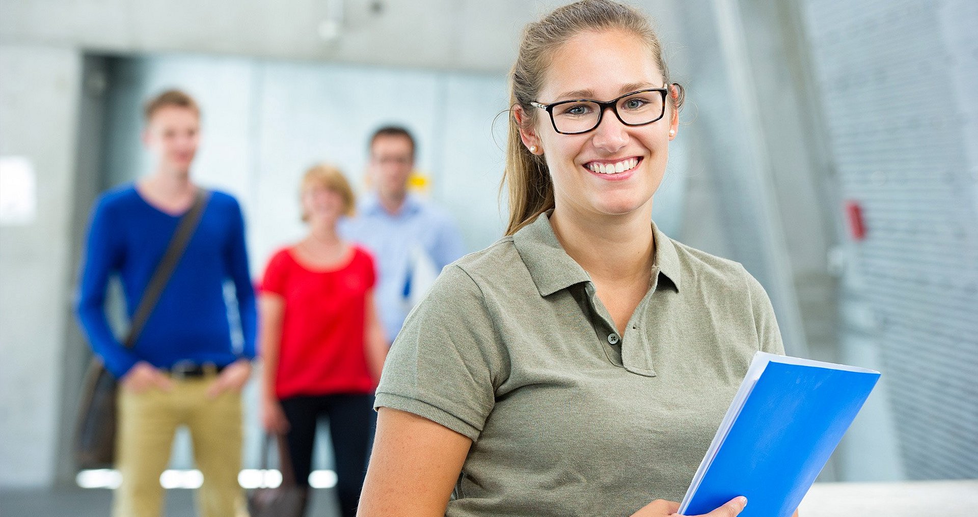 Smiling female student holding a blue folder A female student is standing in the room, smiling and holding a blue folder in her hand, while three other students are visible out of focus in the background.