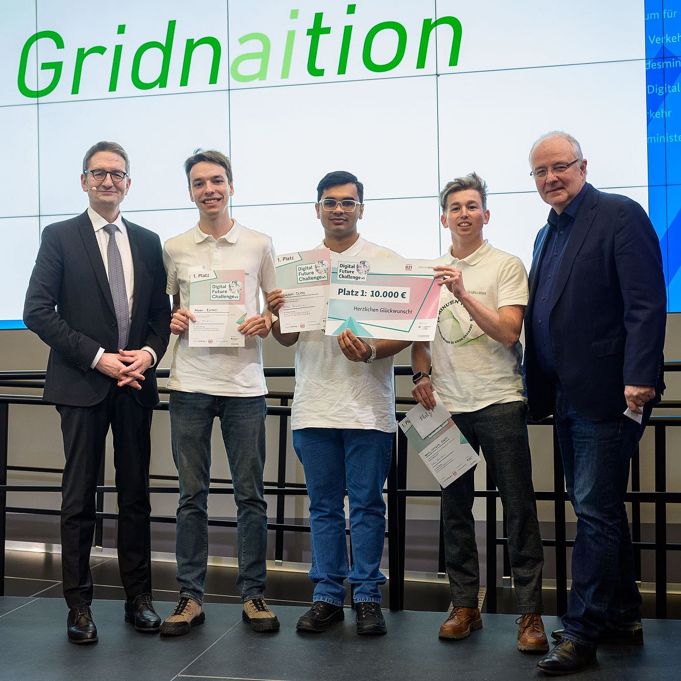 Drei Studierende des Teams „Gridnaition“ auf der Bühne im Bundesministerium für Digitales und Verkehr die Siegerurkunden haltend, links und rechts von ihnen die beiden Laudatoren.