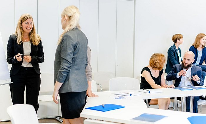 Mehrere Frauen und ein Mann sind in einem Seminarraum. Sie führen eine Gruppenarbeit in jeweils in Zweierteams aus.