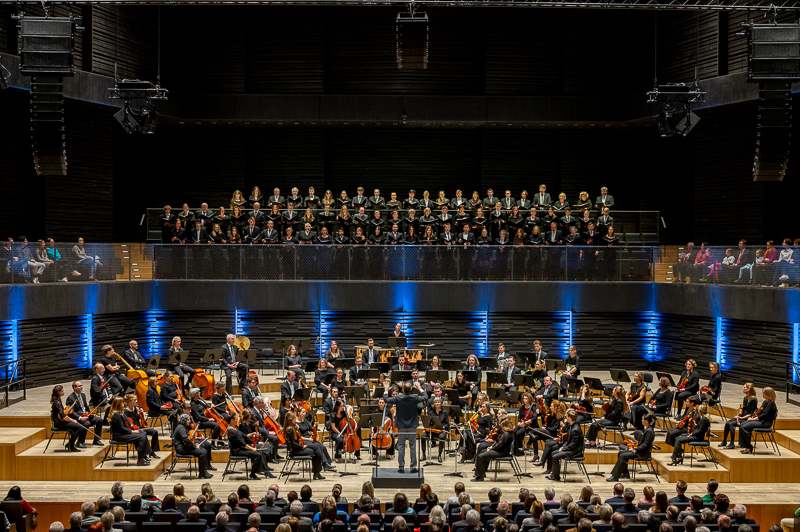 Blick auf die helle Bühne mit dem Orchester vor dunklem Hintergrund und blauen Effektlichtern. Darüber der TUM Chor