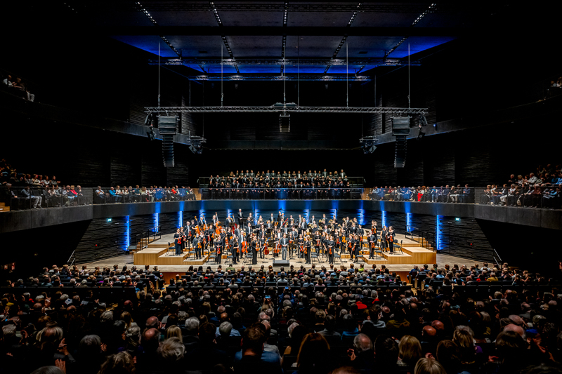 View of the stage from the audience. The orchestra and choir are illuminated - the rest of the hall is dark except for a few blue effect lights