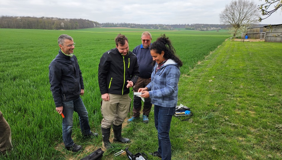 Start-up NutriSen gets the laboratory on the field Eine Frau und drei Männer stehen auf einem Acker. Die Frau zeigt den Männern ein Gerät in ihrer Hand. Es handelt sich um das Gerät, dass das NutriSen-Team zur smarten Düngung entwickelt hat.